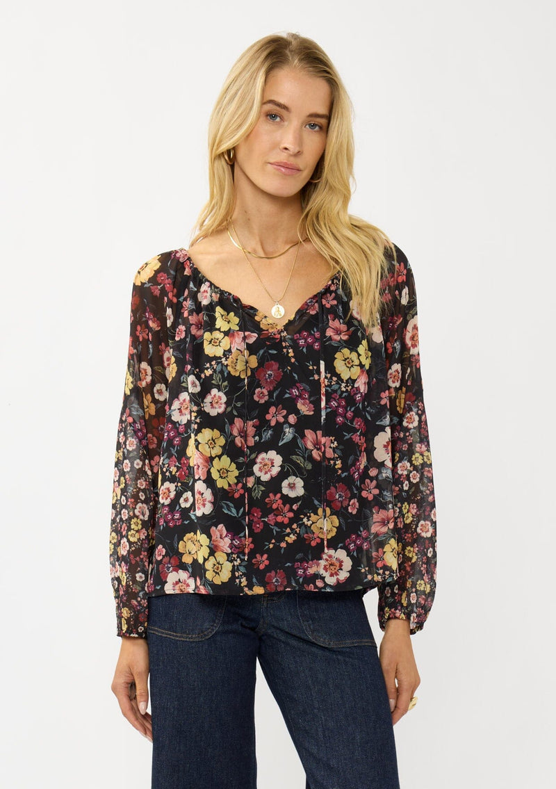 [Color: Black/Rust] A front facing image of a blonde model wearing a black floral sheer chiffon blouse with a split v-neckline with tie detail and long volume sleeves. 
