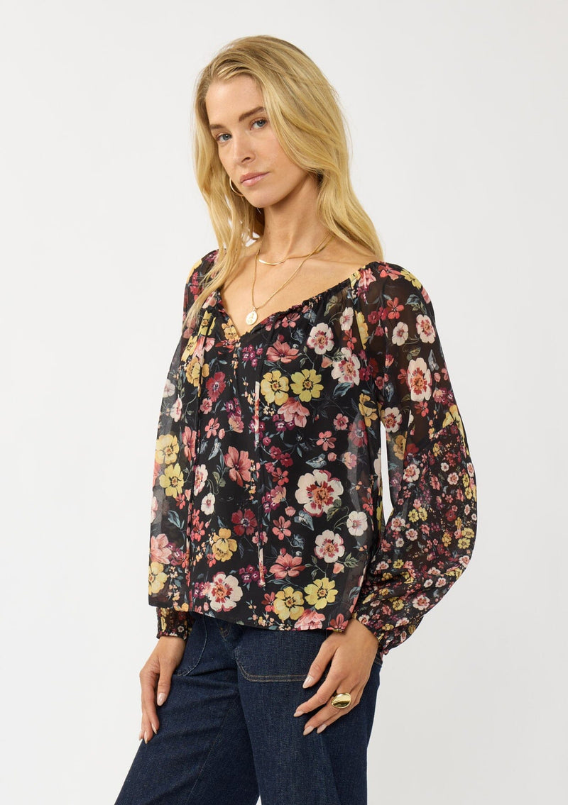 [Color: Black/Rust] A side facing image of a blonde model wearing a black floral sheer chiffon blouse with a split v-neckline with tie detail and long volume sleeves. 