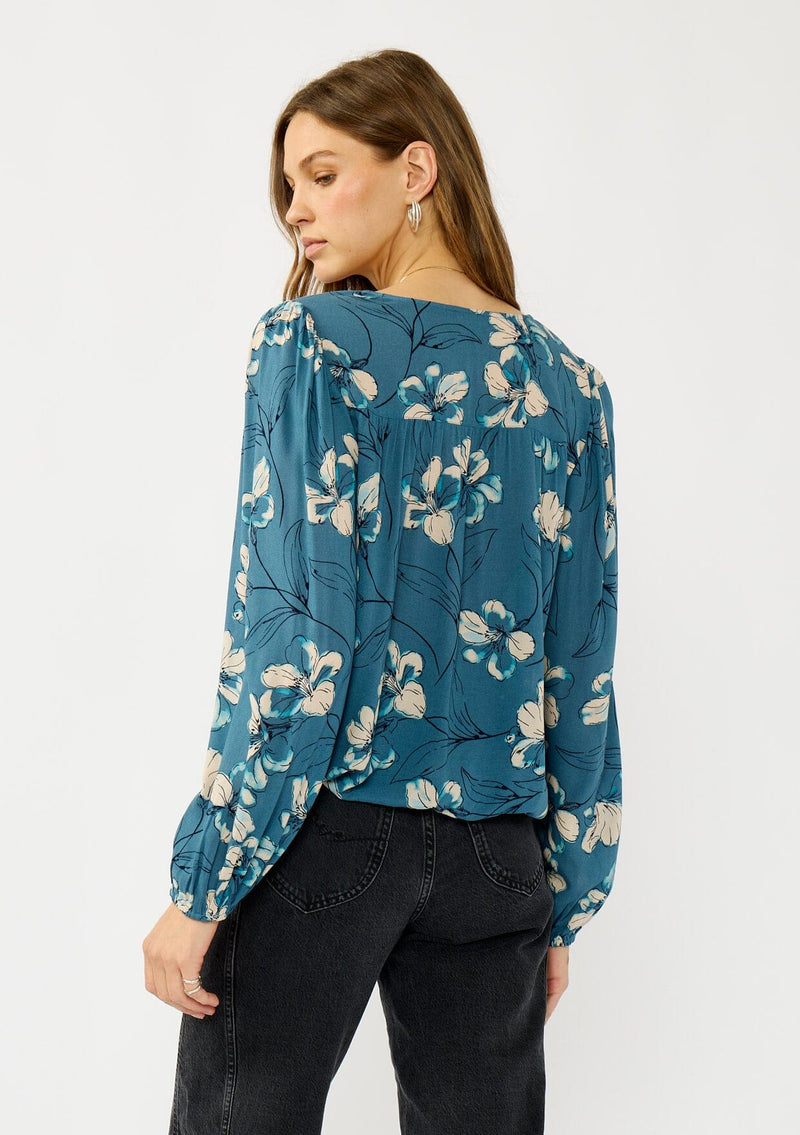 [Color: Teal/Blue] A back facing image of a blonde model wearing a teal floral surplice top with a relaxed fit, long sleeves, front hook and eye, and an elastic hemline. The perfect fall top for casual outings. 