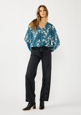 [Color: Teal/Blue] A front facing image of a blonde model wearing a teal floral surplice top with a relaxed fit, long sleeves, front hook and eye, and an elastic hemline. The perfect fall top for casual outings. 