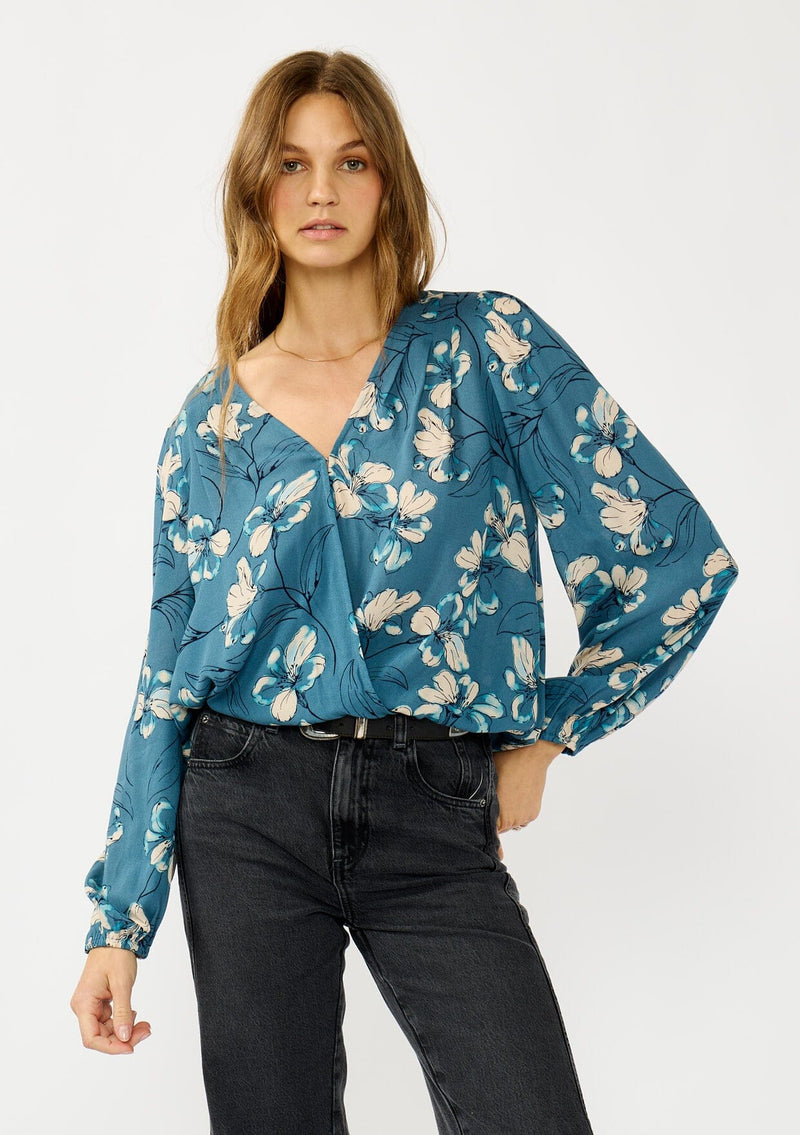 [Color: Teal/Blue] A front facing image of a blonde model wearing a teal floral surplice top with a relaxed fit, long sleeves, front hook and eye, and an elastic hemline. The perfect fall top for casual outings. 