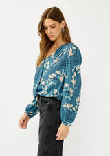 [Color: Teal/Blue] A side facing image of a blonde model wearing a teal floral surplice top with a relaxed fit, long sleeves, front hook and eye, and an elastic hemline. The perfect fall top for casual outings. 