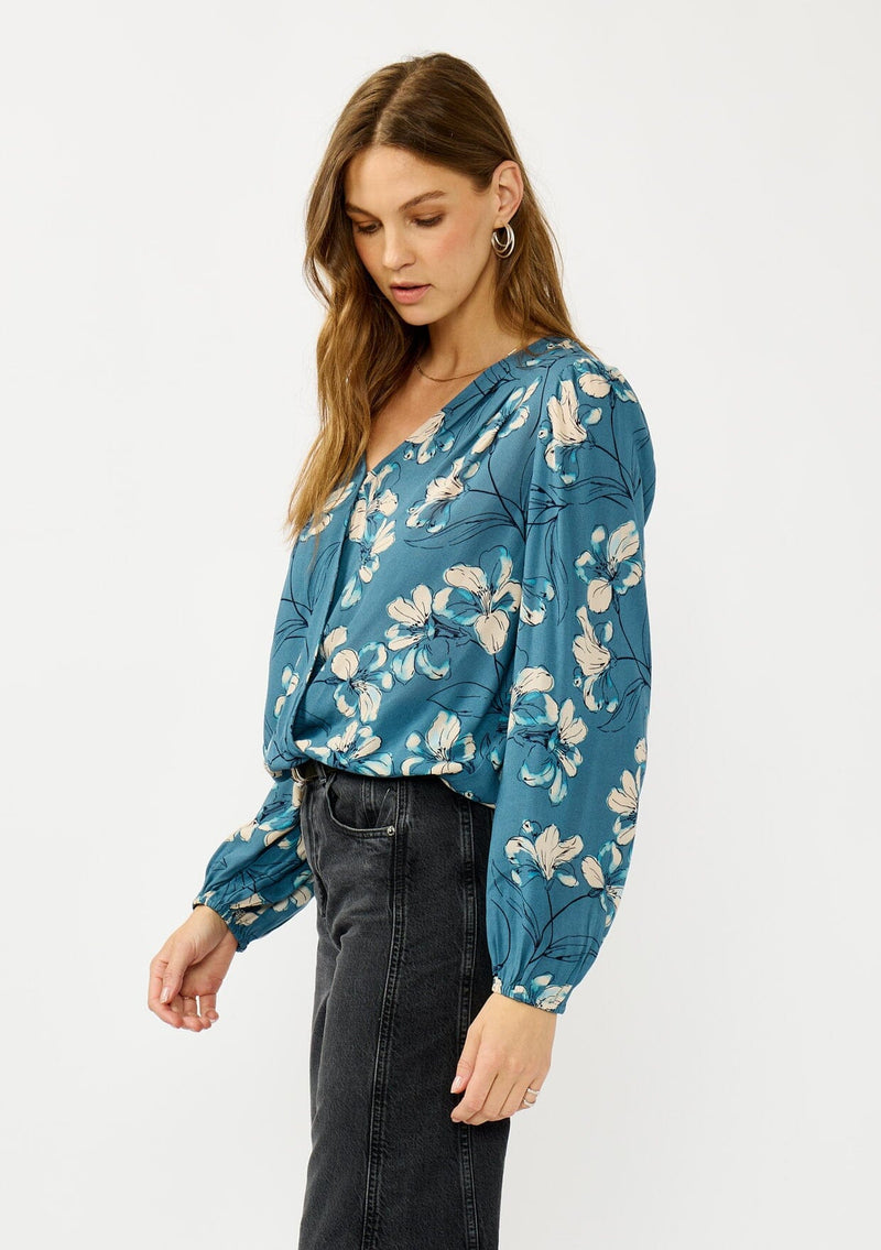 [Color: Teal/Blue] A side facing image of a blonde model wearing a teal floral surplice top with a relaxed fit, long sleeves, front hook and eye, and an elastic hemline. The perfect fall top for casual outings. 