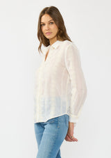 [Color: White/Natural] A side facing image of a brunette model wearing a white embroidered shirt with a button front and collar. 