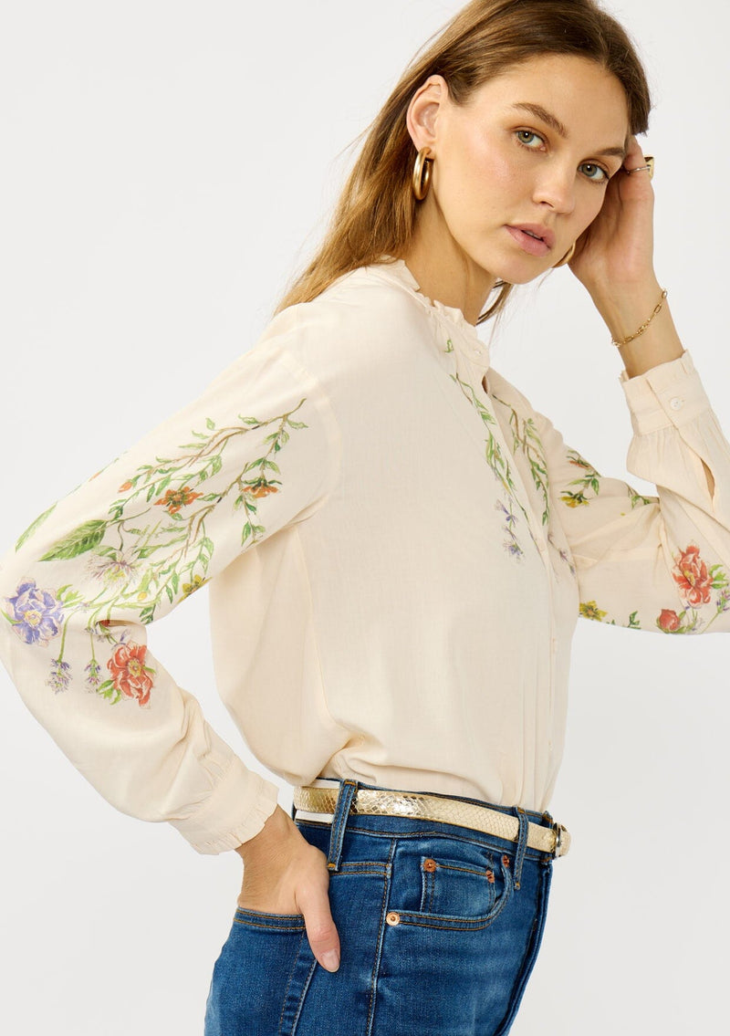 [Color: Stone/Lavender] A side facing image of a blonde model wearing a cream floral blouse with a button front and ruffle trims. A garden style top for special occasions. 