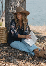 [Color: Eclipse/Coral] A sitting image of a blonde model by a lake wearing a dark blue embroidered blouse with 3/4 length sleeves,  a split v-neckline with tie details, and lattice trim accents. 