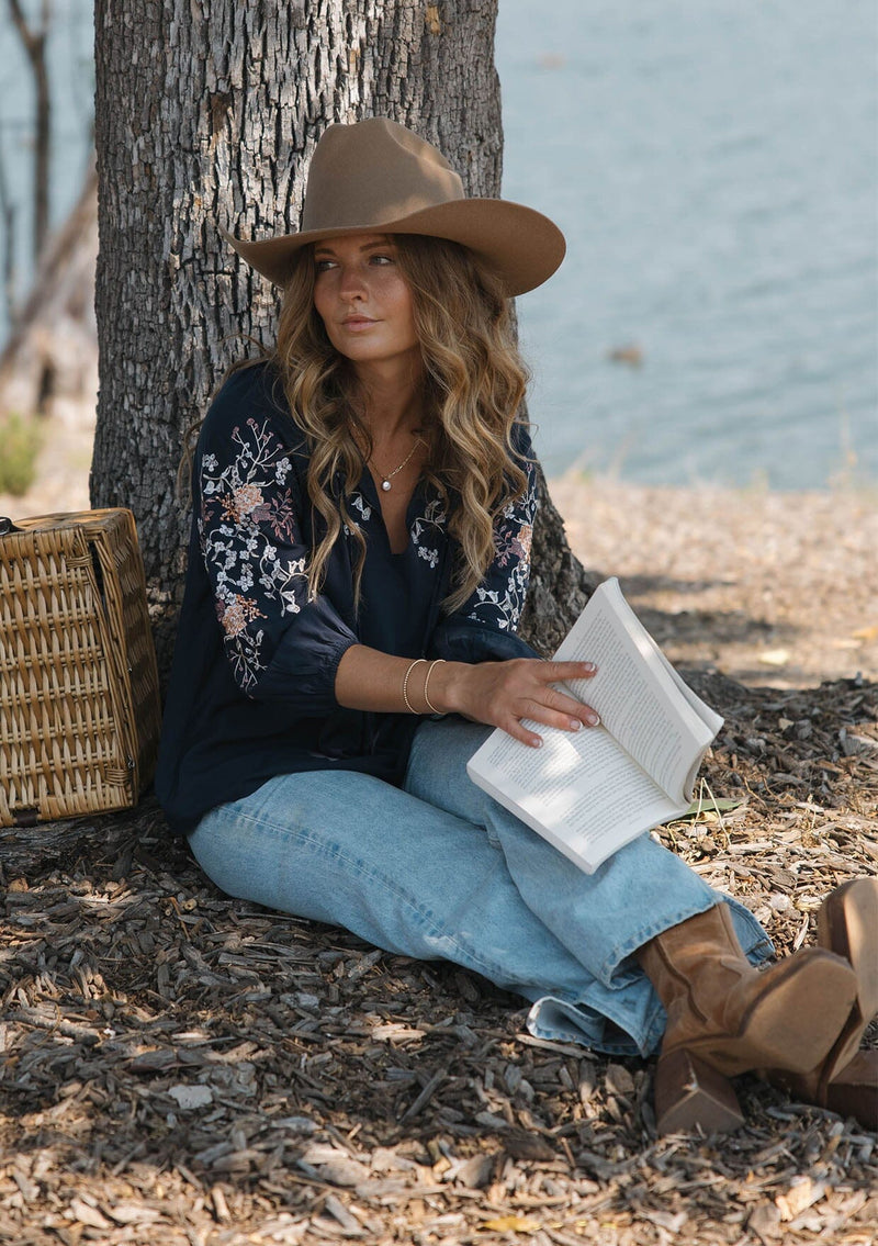 [Color: Eclipse/Coral] A sitting image of a blonde model by a lake wearing a dark blue embroidered blouse with 3/4 length sleeves,  a split v-neckline with tie details, and lattice trim accents. 