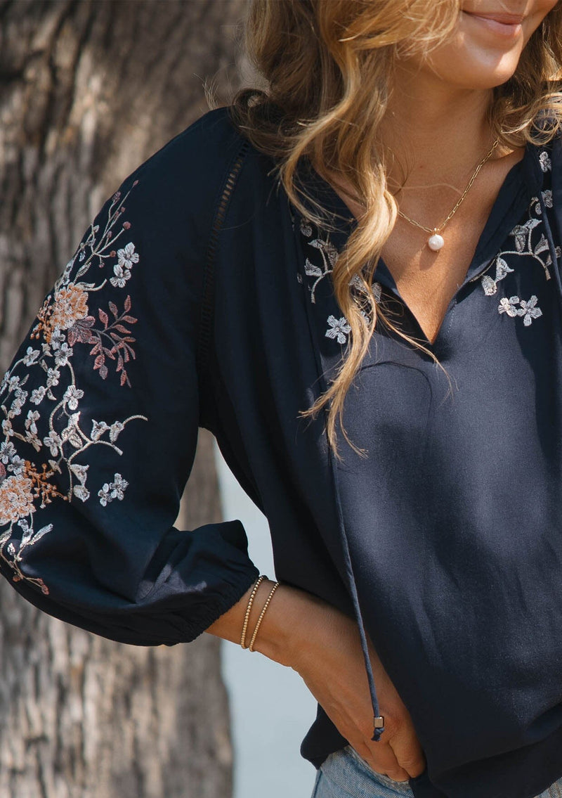 [Color: Eclipse/Coral] A detail image of a blonde model by a lake wearing a dark blue embroidered blouse with 3/4 length sleeves,  a split v-neckline with tie details, and lattice trim accents. 