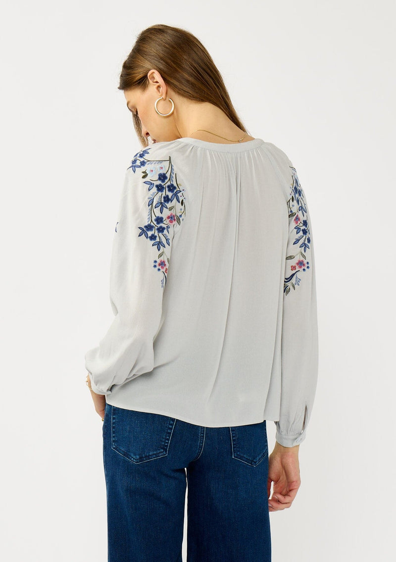 [Color: Light Grey/Navy] A back facing image of a blonde model wearing a light grey blue blouse with floral embroidery, a split v-neckline, and long sleeves with cuffs.  
