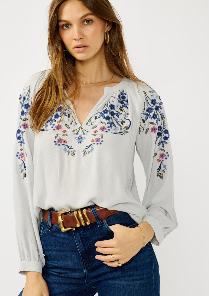 [Color: Light Grey/Navy] A front facing image of a blonde model wearing a light grey blue blouse with floral embroidery, a split v-neckline, and long sleeves with cuffs.  