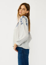 [Color: Light Grey/Navy] A side facing image of a blonde model wearing a light grey blue blouse with floral embroidery, a split v-neckline, and long sleeves with cuffs.  