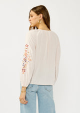 [Color: Pale Rose] A back facing image of a blonde model wearing a light pink blouse with floral embroidery, lace accents, and a flattering split v-neckline. Effortlessly boho and feminine for casual outings. 