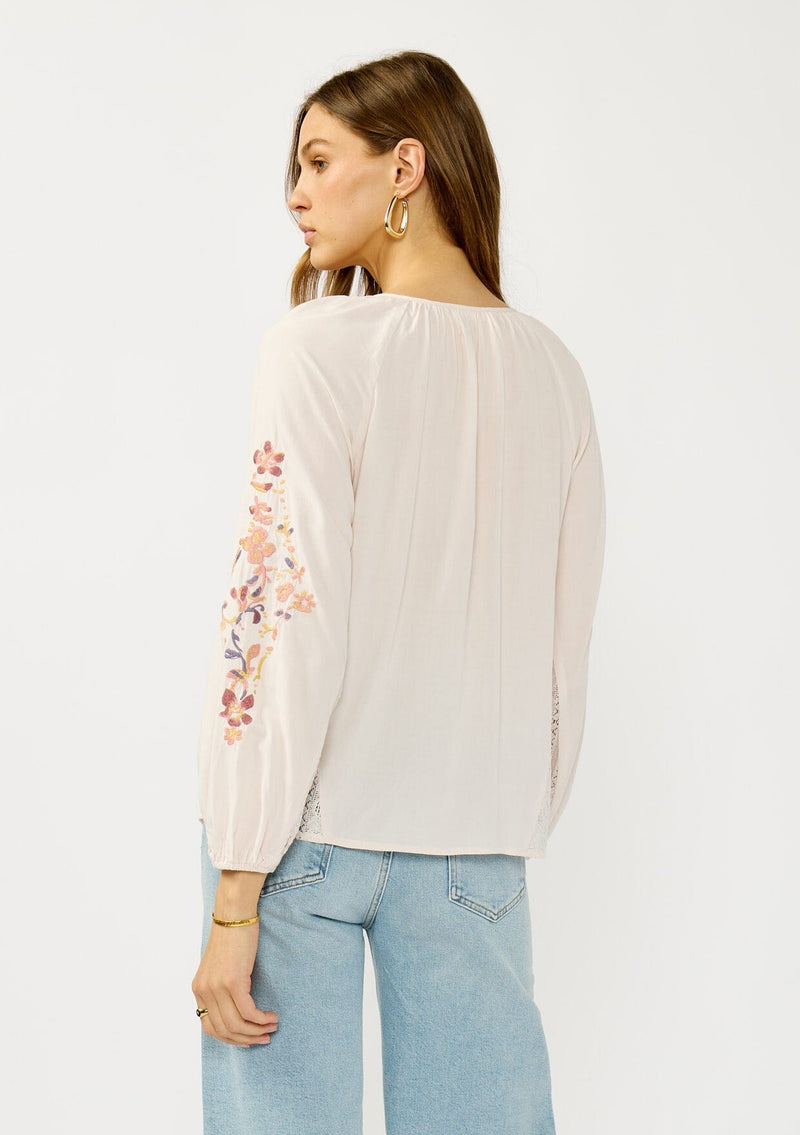 [Color: Pale Rose] A back facing image of a blonde model wearing a light pink blouse with floral embroidery, lace accents, and a flattering split v-neckline. Effortlessly boho and feminine for casual outings. 