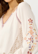[Color: Pale Rose] A detail image of a blonde model wearing a light pink blouse with floral embroidery, lace accents, and a flattering split v-neckline. Effortlessly boho and feminine for casual outings. 