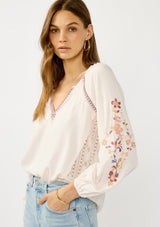 [Color: Pale Rose] A front facing image of a blonde model wearing a light pink blouse with floral embroidery, lace accents, and a flattering split v-neckline. Effortlessly boho and feminine for casual outings. 