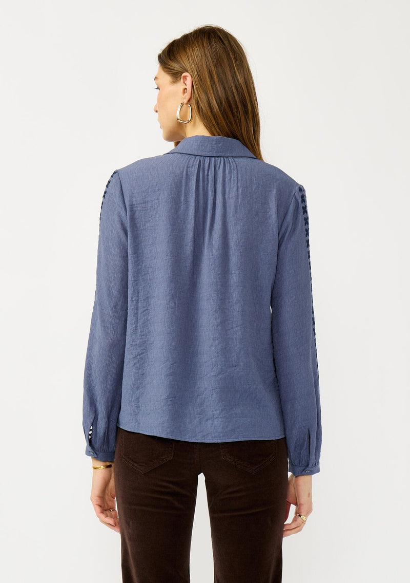 [Color: Denim] A back facing image of a blonde model wearing a blue button up blouse with embroidered details, a collared neckline, and button cuffs. A casual yet elegant top for the fall season. 