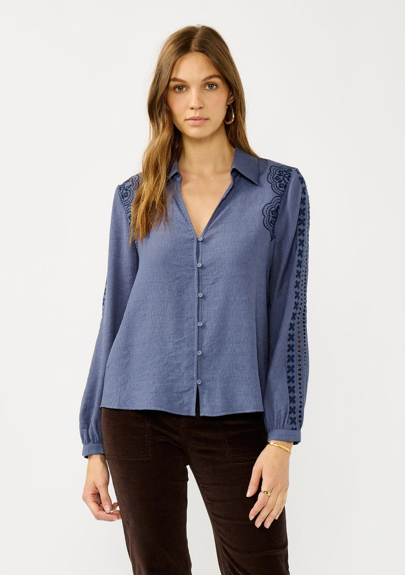 [Color: Denim] A front facing image of a blonde model wearing a blue button up blouse with embroidered details, a collared neckline, and button cuffs. A casual yet elegant top for the fall season. 
