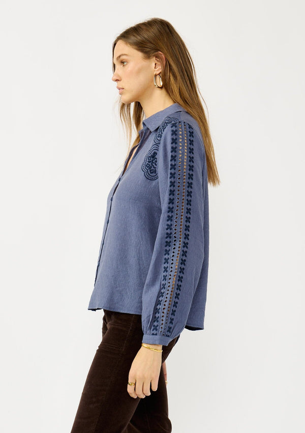 [Color: Denim] A side facing image of a blonde model wearing a blue button up blouse with embroidered details, a collared neckline, and button cuffs. A casual yet elegant top for the fall season. 