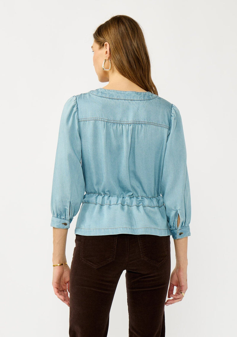 [Color: Light Denim Wash] A back facing image of a blonde model wearing a blue tencel long sleeve blouse with a peplum style waist, drawstring, tie front, v-neckline, and a hidden snap button front. A casual fall blouse. 