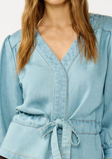 [Color: Light Denim Wash] A detail image of a blonde model wearing a blue tencel long sleeve blouse with a peplum style waist, drawstring, tie front, v-neckline, and a hidden snap button front. A casual fall blouse. 