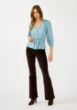 [Color: Light Denim Wash] A front facing image of a blonde model wearing a blue tencel long sleeve blouse with a peplum style waist, drawstring, tie front, v-neckline, and a hidden snap button front. A casual fall blouse. 
