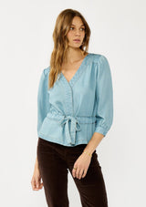 [Color: Light Denim Wash] A front facing image of a blonde model wearing a blue tencel long sleeve blouse with a peplum style waist, drawstring, tie front, v-neckline, and a hidden snap button front. A casual fall blouse. 