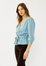 [Color: Light Denim Wash] A side facing image of a blonde model wearing a blue tencel long sleeve blouse with a peplum style waist, drawstring, tie front, v-neckline, and a hidden snap button front. A casual fall blouse. 