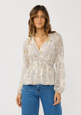 [Color: Natural/Dusty Peach] A front facing image of a brunette model wearing a dotted chiffon peplum blouse with a floral design. Featuring a flattering v-neckline, triple elastic waist, and long sleeves. Accented with metallic clipped details for a touch of sparkle. 