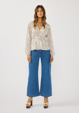 [Color: Natural/Dusty Peach] A front facing image of a brunette model wearing a dotted chiffon peplum blouse with a floral design. Featuring a flattering v-neckline, triple elastic waist, and long sleeves. Accented with metallic clipped details for a touch of sparkle. 