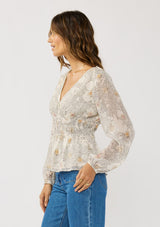 [Color: Natural/Dusty Peach] A side facing image of a brunette model wearing a dotted chiffon peplum blouse with a floral design. Featuring a flattering v-neckline, triple elastic waist, and long sleeves. Accented with metallic clipped details for a touch of sparkle. 