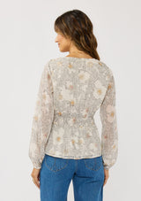 [Color: Natural/Dusty Peach] A back facing image of a brunette model wearing a dotted chiffon peplum blouse with a floral design. Featuring a flattering v-neckline, triple elastic waist, and long sleeves. Accented with metallic clipped details for a touch of sparkle. 
