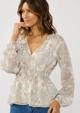 [Color: Natural/Dusty Peach] A front facing image of a brunette model wearing a dotted chiffon peplum blouse with a floral design. Featuring a flattering v-neckline, triple elastic waist, and long sleeves. Accented with metallic clipped details for a touch of sparkle. 