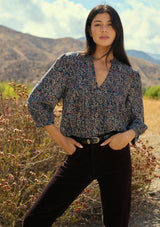 [Color: Navy/Rose] A front facing image of a brunette model wearing a navy floral blouse with 3/4 length sleeves, a split v-neckline, and front pintuck details. A classic boho blouse for casual outings. Woman standing in a natural setting with mountains and plants