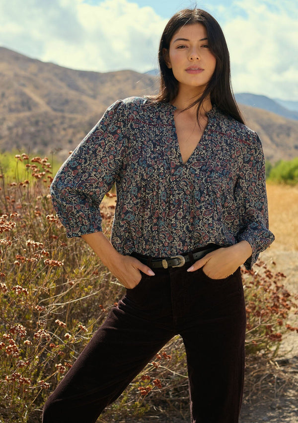 [Color: Navy/Rose] A front facing image of a brunette model wearing a navy floral blouse with 3/4 length sleeves, a split v-neckline, and front pintuck details. A classic boho blouse for casual outings. Woman standing in a natural setting with mountains and plants