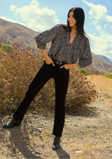 [Color: Navy/Rose] A front facing image of a brunette model wearing a navy floral blouse with 3/4 length sleeves, a split v-neckline, and front pintuck details. A classic boho blouse for casual outings. Woman standing in a natural setting with mountains and plants
