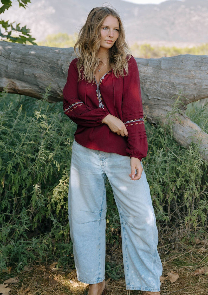 [Color: Berry] A front facing image of a blonde model wearing a burgundy boho blouse with colorful embroidered details, a split v-neckline with ties, and delicate pintuck details along the sleeve. 