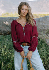 [Color: Berry] A front facing image of a blonde model wearing a burgundy boho blouse with colorful embroidered details, a split v-neckline with ties, and delicate pintuck details along the sleeve. 