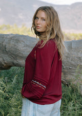 [Color: Berry] A side facing image of a blonde model wearing a burgundy boho blouse with colorful embroidered details, a split v-neckline with ties, and delicate pintuck details along the sleeve. 