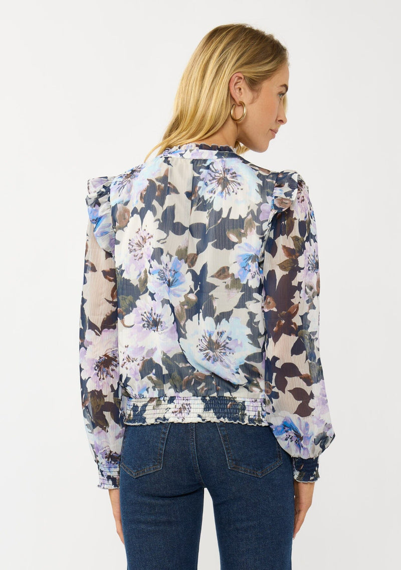 [Color: Grey/Navy] A back facing image of a blonde model wearing a grey and purple floral top with a split v-neckline, smocked waist and cuff, and a ruffle shoulder detail. A chic fall blouse for special occasions. 