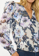 [Color: Grey/Navy] A detail image of a blonde model wearing a grey and purple floral top with a split v-neckline, smocked waist and cuff, and a ruffle shoulder detail. A chic fall blouse for special occasions. 