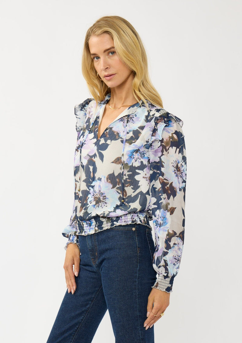 [Color: Grey/Navy] A side facing image of a blonde model wearing a grey and purple floral top with a split v-neckline, smocked waist and cuff, and a ruffle shoulder detail. A chic fall blouse for special occasions. 