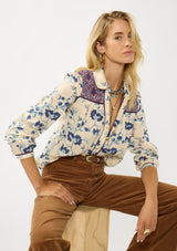 [Color: Cream/Plum] A sitting image of a blonde model wearing a purple and cream floral boho shirt with a classic collar neck and button front. A unique fall top for casual styling. 