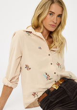 [Color: Vanilla] A sitting image of a blonde model wearing a corduroy shirt with floral embroidery, a button front, and a collar neck. A soft and comfortable shirt for the fall season. 