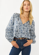 [Color: Blue] A front facing image of a blonde model wearing a blue floral blouse with a subtle sheen. Featuring long sleeves with shirred details, a split v-neckline with tie detail, and a ruffled neckline. 
