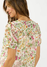[Color: Blush/Rose] A detail image of a brunette model wearing a pink and green floral v neck top with a button front and a ruffle trim accent. 