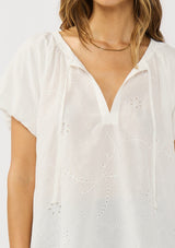 [Color: White] A detail image of a brunette model wearing a white puff sleeve blouse with an eyelet inspired design and a split v-neckline with ties. 