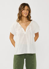 [Color: White] A front facing image of a brunette model wearing a white puff sleeve blouse with an eyelet inspired design and a split v-neckline with ties. 