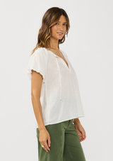 [Color: White] A side facing image of a brunette model wearing a white puff sleeve blouse with an eyelet inspired design and a split v-neckline with ties. 