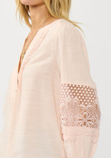[Color: Blush] A side facing image of a blonde model wearing a pink blouse with a button front, split v-neckline, pintuck details, and a lace applique on the sleeves. A warm weather style part of the resort collection. 