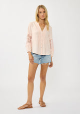 [Color: Blush] A front facing image of a blonde model wearing a pink blouse with a button front, split v-neckline, pintuck details, and a lace applique on the sleeves. A warm weather style part of the resort collection. 